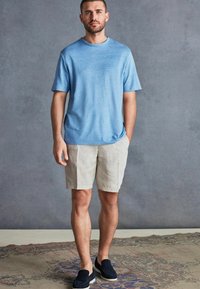 Tricou bleu deschis cu mânecă scurtă, asortat cu pantaloni scurți din in bej și pantofi slip-on bleumarin, plasat pe un fundal neutru.