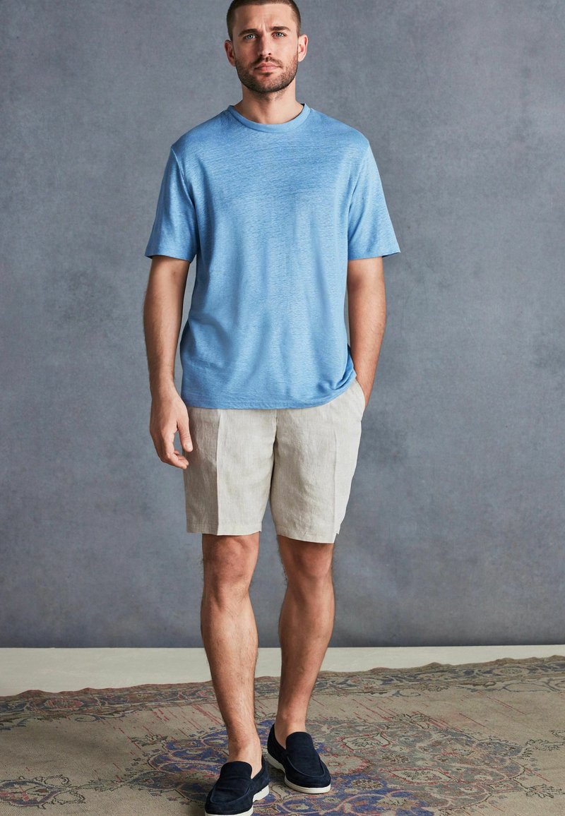 Tricou bleu deschis cu mânecă scurtă, asortat cu pantaloni scurți din in bej și pantofi slip-on bleumarin, plasat pe un fundal neutru.