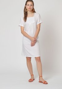 Vestido de algodón blanco con mangas cortas, escote cuadrado con borde de encaje y ajuste relajado. Combinado con sandalias naranjas de tira.