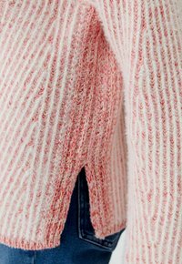 Nahaufnahme eines rosa-weißen, gerippten Strickpullovers mit seitlichem Schlitz über blauen Jeans.