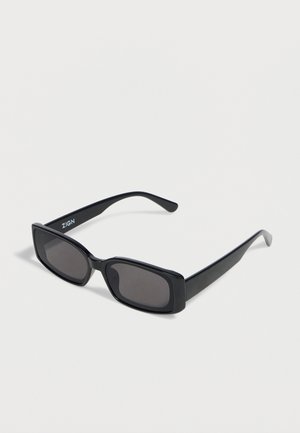 Lunettes de soleil rectangulaires noires avec verres foncés et montures épaisses, présentées en angle sur un fond blanc.