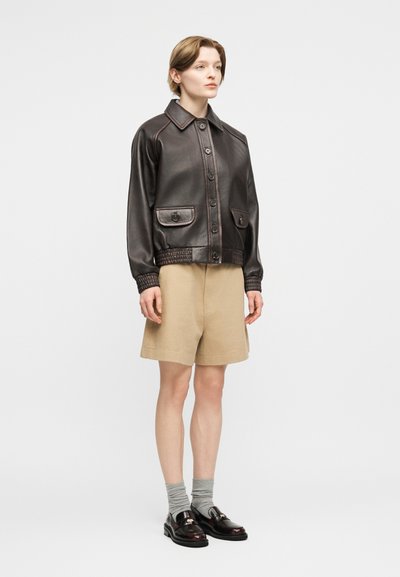 Άτομο που φοράει σκουρόκαφέ δερμάτινο μπουφάν με κουμπιά, μπεζ σορτς μέχρι το γόνατο, γκρι κάλτσες και σκουρόκαφέ μοκασίνια, στέκεται μπροστά σε λευκό φόντο.