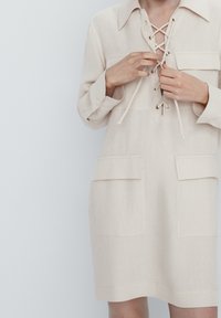 Beiges Leinenkleid mit einem Kragen, vorderem Schnürdesign und zwei aufgesetzten Taschen auf der Brust. Verfügt über umgeschlagene Ärmel und einen geraden Schnitt.