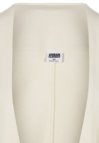Cream-colored garment with a textured fabric, wide collar, and a tag labeled "URBAN CLASSICS XS." Simple and minimal design.