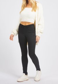 Černé skinny džíny v kombinaci s bílým krátkým pleteným svetrem a krémovým kardiganem. Model má na sobě bílé tenisky, což podtrhuje neformální outfit.