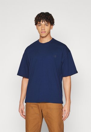 Jaunas vyras su trumpais garbanotais plaukais, dėvintis laisvą tamsiai mėlyną marškinėlį ir rudus kelnes, stovintis prie vienodo šviesaus fono.