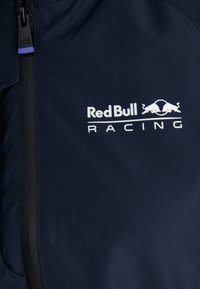 Chaqueta azul marino con un destacado logo blanco de "Red Bull Racing". Tela suave con un diseño elegante y cremallera negra en contraste.