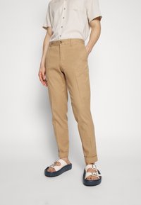 Beige, tapered byxor med flat front, bälteshällor och uppvikta ärmar, i kombination med vita sandaler med mörk sula och silverdetaljer.