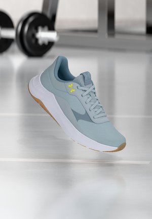 Svetlomodrá športová obuv s bielou podrážkou a gumovým vzorom, označená žltým logom Under Armour, umiestnená na odrazovej podlahe telocvične s činmi v pozadí.