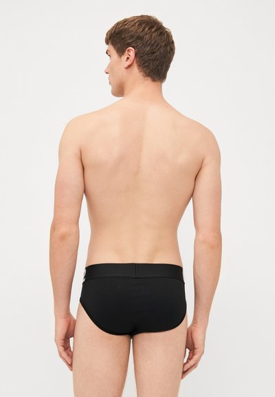 Schwarze Schwimmslips mit glatter Textur, ausgestattet mit einem breiten elastischen Bund und einem tiefen Schnitt, von hinten gesehen.