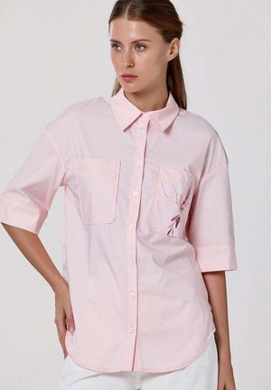 Femme portant une chemise rose clair boutonnée avec deux poches à l’avant, dont une ornée d’un petit motif floral violet, et un pantalon blanc.