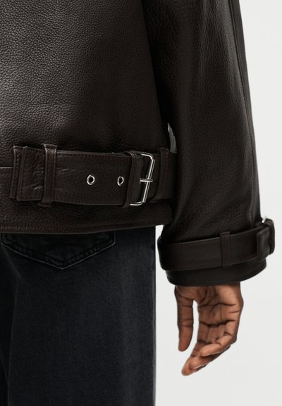 Κοντινό πλάνο σκούρου καφέ υφασμάτινου δερμάτινου μπουφάν με ρυθμιζόμενη ζώνη και ιμάντες στο μανίκι, φορεμένο πάνω από μαύρο τζιν.