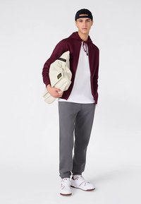 Sudadera con cremallera color marrón oscuro, camiseta blanca, pantalones deportivos grises, sosteniendo una bolsa crema. Zapatos blancos con detalles rojos. Conjunto de ropa deportiva simple y casual.