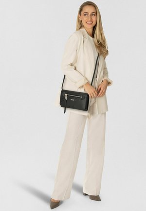 Schwarze Umhängetasche, vorne mit Reißverschlusstasche, Logodetail, glatte Textur, kombiniert mit einem beigen Anzug und grauen High Heels, vor neutralem Hintergrund.