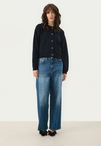 Navy knitted cardigan with a round neckline, buttons, and pockets, paired with light blue wide-leg jeans and black bow-accented heels.