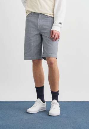 Hombre que lleva pantalones cortos a rayas azules y blancas hasta la rodilla, calcetines negros, zapatillas blancas y una camiseta de manga larga color crema con un diseño de araña en la manga.