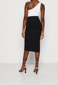 Kvinna klädd i en svart högmidjad midikjol, vit off-the-shoulder topp och svarta snörda högklackade sandaler vänd bort mot en enfärgad bakgrund.