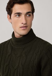 Ung man med kort, lockigt hår iklädd en mörkgrön kabelstickad polotröja, som tittar åt sidan mot en neutral bakgrund.