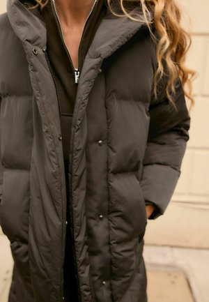 Manteau matelassé gris foncé avec fermeture éclair à l'avant, grand col et poches avant, avec une texture matelassée et des détails à bouton-pression.