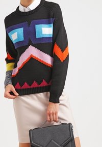 Schwarzer gestrickter Pullover mit geometrischen, mehrfarbigen Mustern, mit Rundhalsausschnitt, kombiniert mit einem hellen Kleid und einer schwarzen Handtasche.