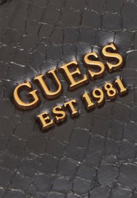 Sort tekstureret læder med hævede gyldne bogstaver "GUESS EST 1981", der fremhæver en glansfuld finish og et detaljeret overflademønster.