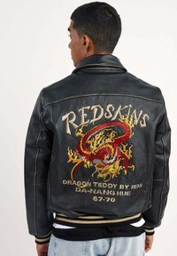 Veste en cuir noir avec un grand design de dragon rouge brodé et le texte "REDSKINS". Les détails comprennent un col et un ourlet tricotés.