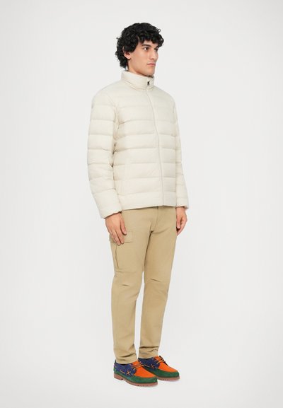 Giacca imbottita beige con cuciture verticali, colletto alto e chiusura con zip, abbinata a pantaloni cargo color sabbia e scarpe in suede multicolori.