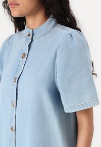 Lyseblå denimskjorte med korte ermer, høy krage og knappeåpning. Har lett puff ved skulderen og synlige sømdetaljer.