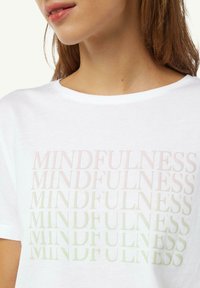 Biała koszulka z powtarzającym się napisem "MINDFULNESS" w gradiencie od jasnego różu do zieleni. Okrągły dekolt i miękki materiał bawełniany.
