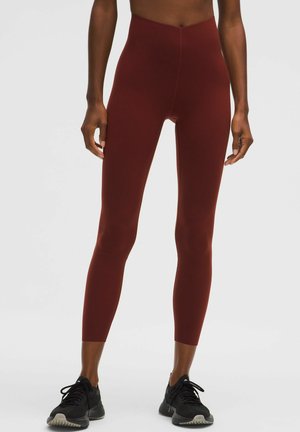 Højtsiddende, tætsiddende leggings i dyb rød, lavet af glat, strækbart stof. Båret med sorte træningssko. Ingen synlige logoer.