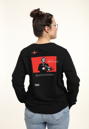 Černá mikina s grafikou na zádech, která má červené pozadí, postavu držící zbraň a japonský text. Logo "SCARFACE" je viditelné.
