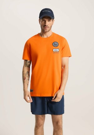 Pomarańczowa bawełniana koszulka z okrągłym dekoltem, krótkimi rękawami i haftowanymi naszywkami. Noszona z ciemnoniebieskimi szortami. Prosty i skromny design.