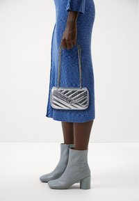 Borsa a tracolla trapuntata argento con manico a catena, abbinata a un vestito blu strutturato e stivaletti alla caviglia grigio chiaro, con tacco blocco.