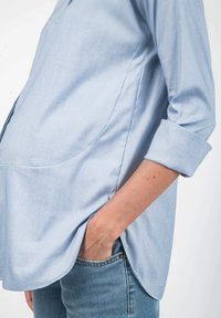 Helles blaues Langarmhemd mit abgerundetem Saum, aus weichem Stoff. Verfügt über Seitentaschen und hochgekrempelte Ärmel.
