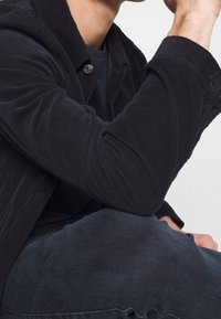 Svart kordflodjacka med texturerade ränder, med en knappstängning. Kombinationen med mörka jeansbyxor ger en avslappnad passform och rena linjer.