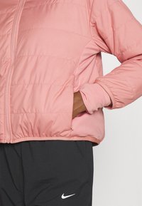 Doudoune rose clair avec des coutures horizontales, des poignets élastiques et des poches latérales. Portée sur un pantalon noir avec un logo blanc.
