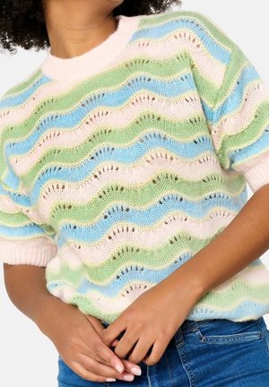 Femme portant un pull tricoté pastel avec des rayures ondulées vertes, bleues et roses, associée à un jean bleu, les mains jointes à la taille.