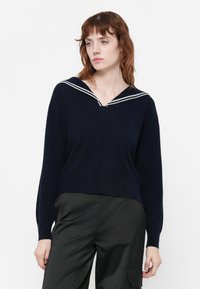 Femme aux cheveux roux portant un pull bleu marine avec des rayures blanches au col et un pantalon cargo noir, se tenant contre un fond blanc.