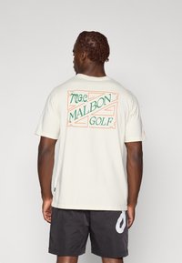 Kremo spalvos medvilnės marškinėliai su spalvinga „M.G.C. MALBON GOLF“ grafika nugaros pusėje, trumpomis rankovėmis ir laisvu siluetu.