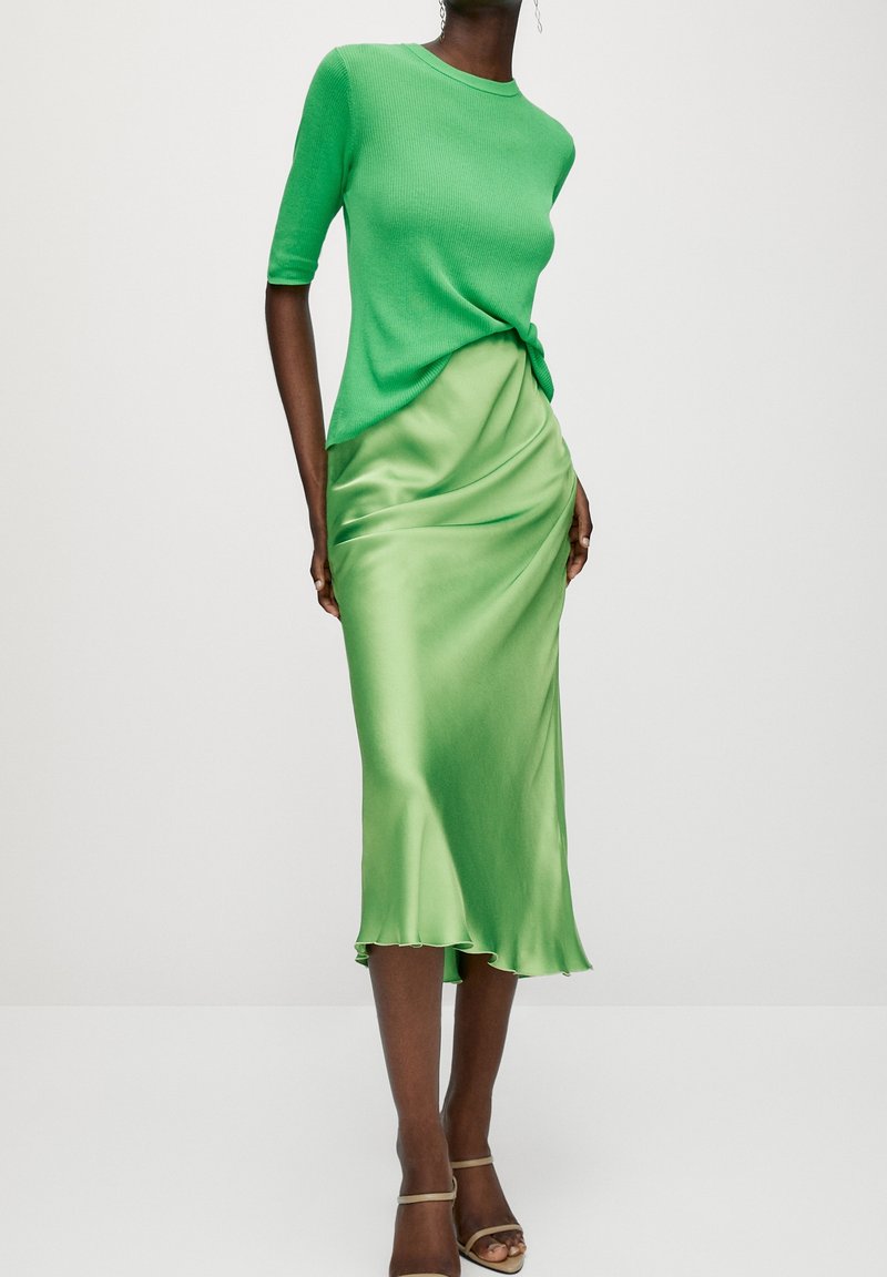 Femme portant un haut à manches courtes vert vif noué à la taille et une jupe midi en satin vert clair brillant, avec des talons à brides à la cheville.