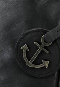Zwart leermateriaal heeft een ronde patch met een messing ankerembleem, gestikt met een zichtbare zwarte draaddetail.