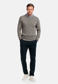 Grijze gebreide trui met een textuurpatroon, gedragen over een blauw geruite blouse, gecombineerd met donkere broek en lichte sneakers.