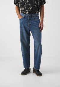 Vaqueros de denim azul con corte recto, que presentan un diseño clásico de cinco bolsillos y un ligero desgaste. Los lleva puestos con zapatillas negras y una camisa de estampado.