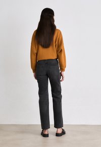 Suéter mostaza, jeans negros de pierna recta, textura suave, detalle de costura mínima, combinado con zapatos negros de tipo slip-on, visto desde la parte de atrás.
