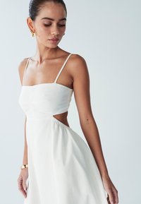 Hvid, ærmeløs kjole med tætsiddende korset og udskæringsdetalje. Flydende nederdel med samlet struktur. Enkel, minimalistisk design.