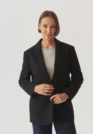 Black blazer with notched lapels, tailored fit, and smooth texture; worn over a gray shirt, paired with dark denim pants.
