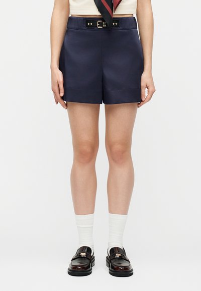 Γυναίκα που φοράει ναυτική tailored σορτς με ζώνη, λευκές κάλτσες και σκούρα γυαλισμένα μοκασίνια, στέκεται μπροστά σε λευκό φόντο.