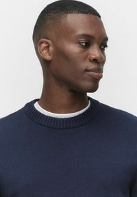 Ung man med kort hår iförd en marinblå tröja över en vit skjorta, som tittar åt höger mot en ljus, enkel bakgrund.