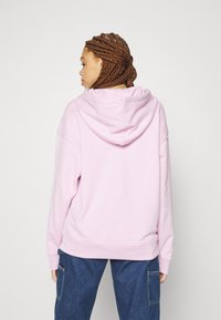 Rosa hettegenser med avslappet passform, med stor hette og ribbestrikket mansjetter. Mykt stoff og en jevn tekstur, brukt sammen med blå denim bukser.