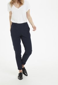 Pantalons stretch navy avec une coupe fuselée et une taille élastique, associés à un t-shirt blanc à col en V et des chaussures noires à lacets.